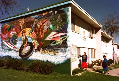 Two women knock on a door of a building with a mural painted on its side. 