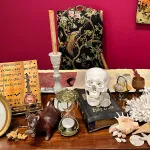 View of an art installation composed of various objects, including a candle, skull, hourglass, bible, shells, and photograph.