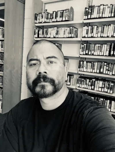 A Latinx man in glasses posed before a bookshelf.