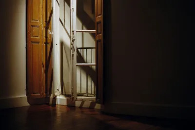 A tall balcony window with indoor wood shutters is a jar in a Parisian room. It is illuminated by a spotlight. 