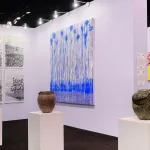 View of paintings hanging on walls in an art fair booth with sculptures on plinths in front.