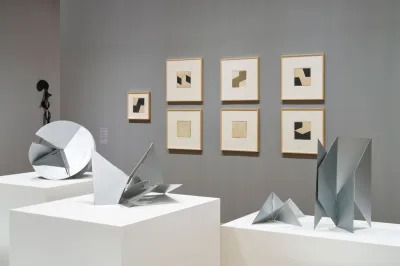 Metal sculptures that can be folded in multiple ways on plinths with similar geometric drawings hanging on the wall behind it. 