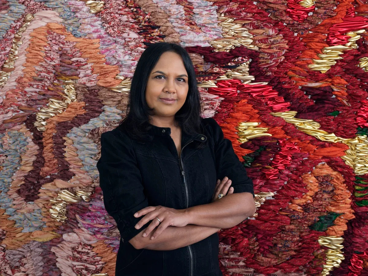 A photograph of a medium-skinned woman with black hair, shown from the waist up. She is wearing a black zip-up jacket and has her arms crossed in front of her. She stands in front of a large-scale abstract tapestry made of red, orange, pink, and gold fabric.