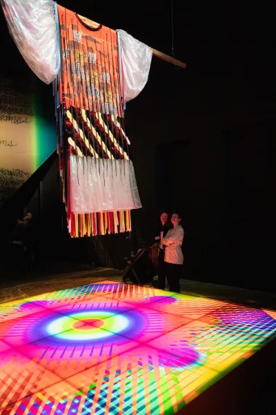 Two viewers peering up at a ceremonial garment, with a lit-up floor by their feet.