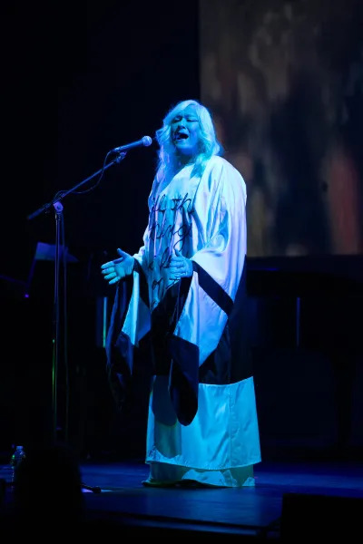 A figure in a white garment with white hair, singing.