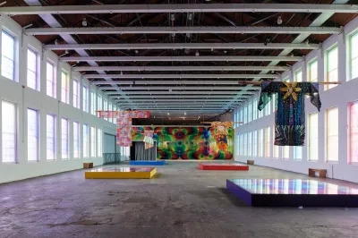 A large room with light-filled windows and three hanging garments over plinths on the floor.