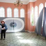 A woman with reddish hair standing amid giant abstract paintings in shades of grey. One leaned against a windowed wall features rings of white and grey that meet at a central cleavage.