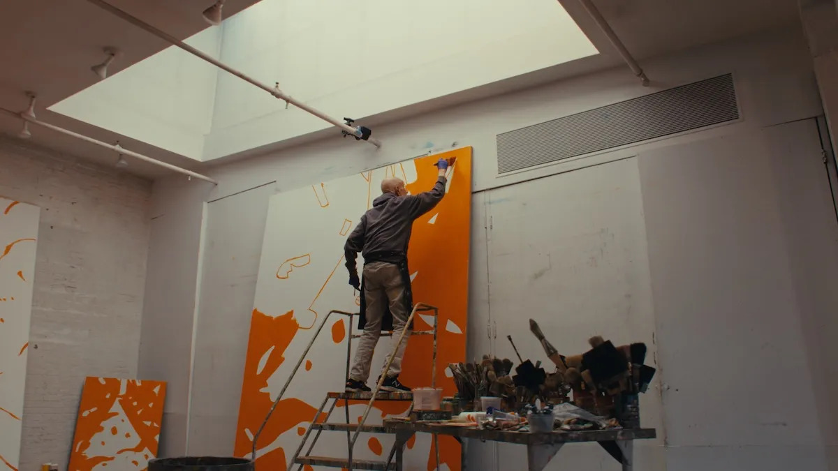 An image of a man standing on a ladder near the top of a large orange and white painting.