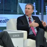 A middle-aged man in a dark suit, blue shirt, and reddish tie sits in a large white chair. He holds his hands in front of himself. He seems to be speaking to someone.