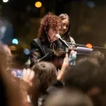 A white woman in a black jacket speaking at a podium before a crowd.