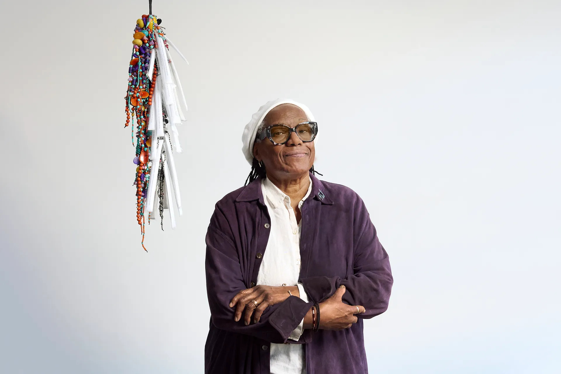 A Black woman holding her arms across her chest. Behind her, there is a hanging sculpture composed of strung-up beads.