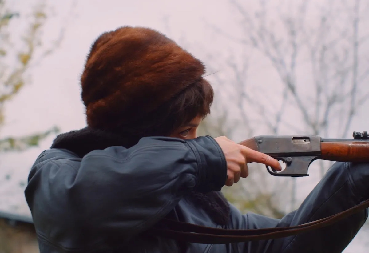 A woman firing a gun seen from the side. She wears a bushy brown hat and a blue shirt.