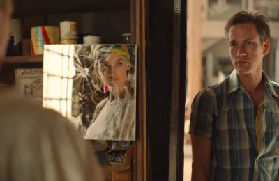 A woman looking in a broken mirror while a man in a plaid shirt looks at her.