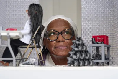 A Black woman wearing glasses looking at an installation showing a Black female doll sitting at a table in a folding chair.