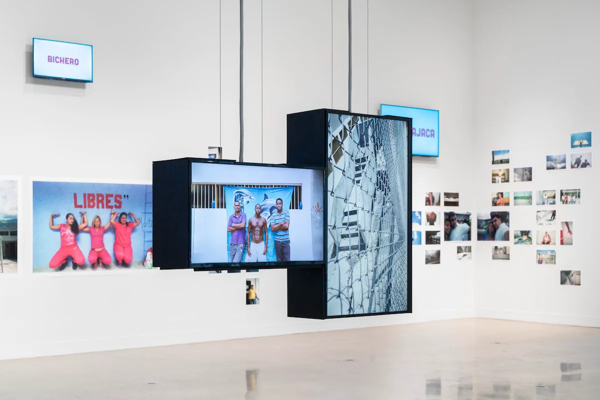 A white-walled gallery with many photos of incarcerated subjects. In the center of the image hang two lightboxes with images.