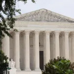 A grand, white, classical building with columns is seen from a distance. Trees loom in the foreground in this color photographs.