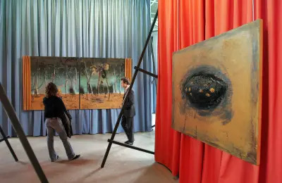 People wandering through an art exhibition whose walls are covered in blue and red curtains. There are paintings hanging over the curtains.