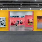 A brightly colored yellow and pink booth at an art fair with many artworks hanging on the walls.