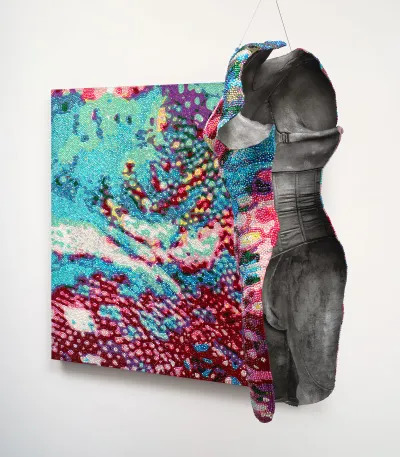 A sculpture hangs in front of a painting. One side of the sculpture and the painting have a similar camouflaged, bejewled pattern. The back of the sculpture has a cast of a body in a corset.  