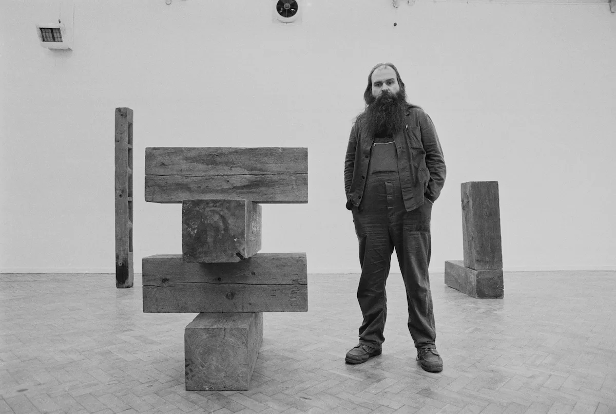 A white man in jeans and a jacket standing beside sculptures composed of wood.