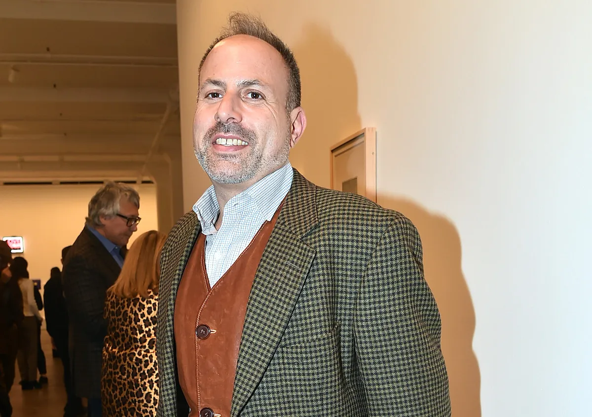 A man in a leather vest and a crosshatched suit jacket smiling in an art gallery.