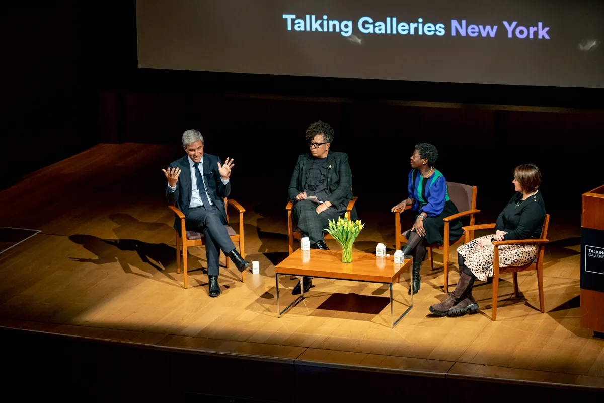 A group of people on a stage sitting around a table with flowers on it and talking.