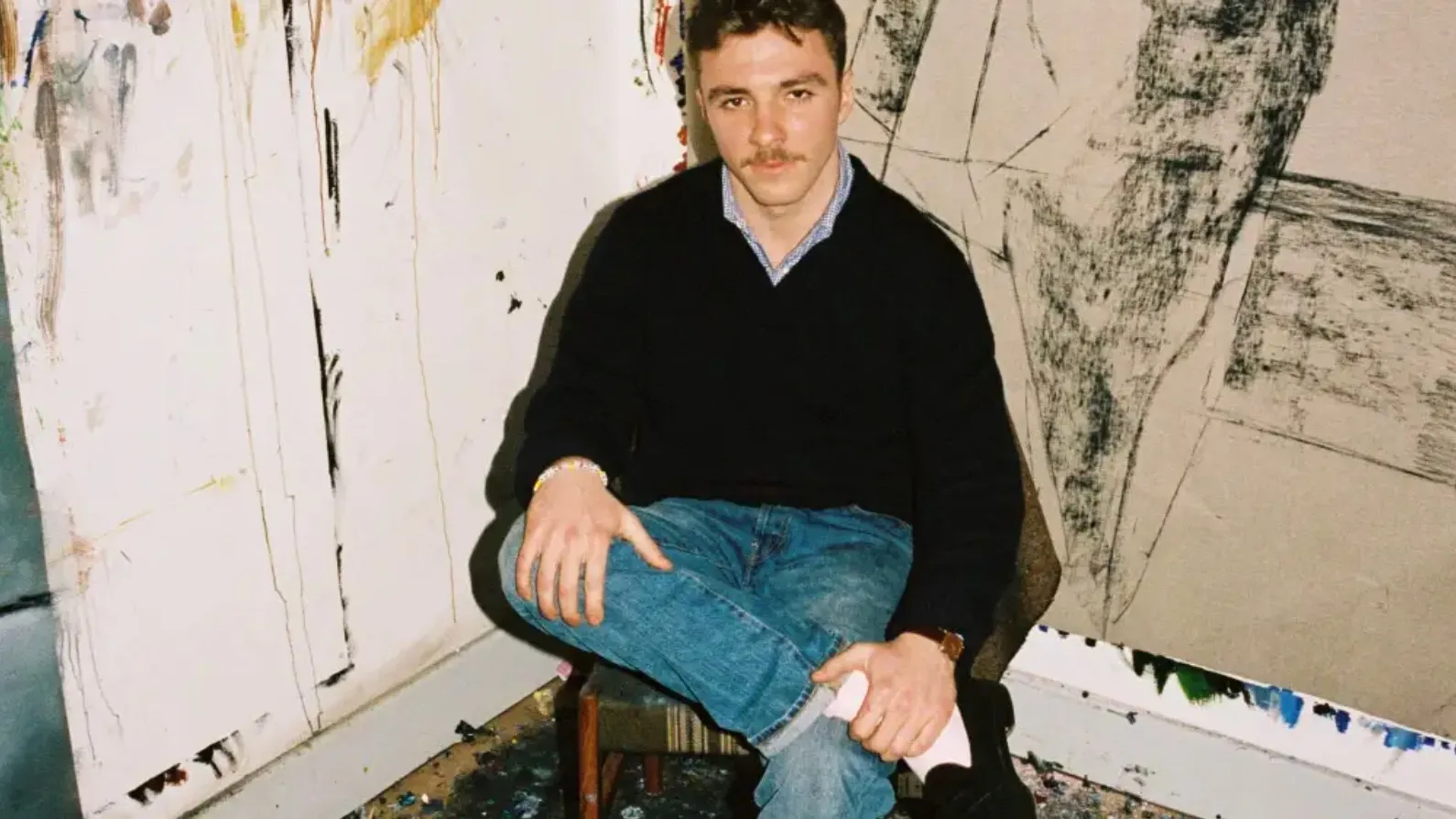A white man in blue jeans sitting cross-legged on a chair between two paintings.