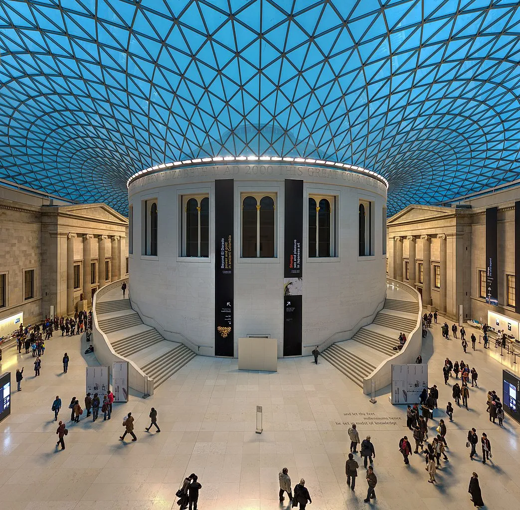 In a color photo, a tan cylindrical structure is seen under a blue, curving canopy. Inside this covered space, many people roam around. Neoclassical buildings are in back.