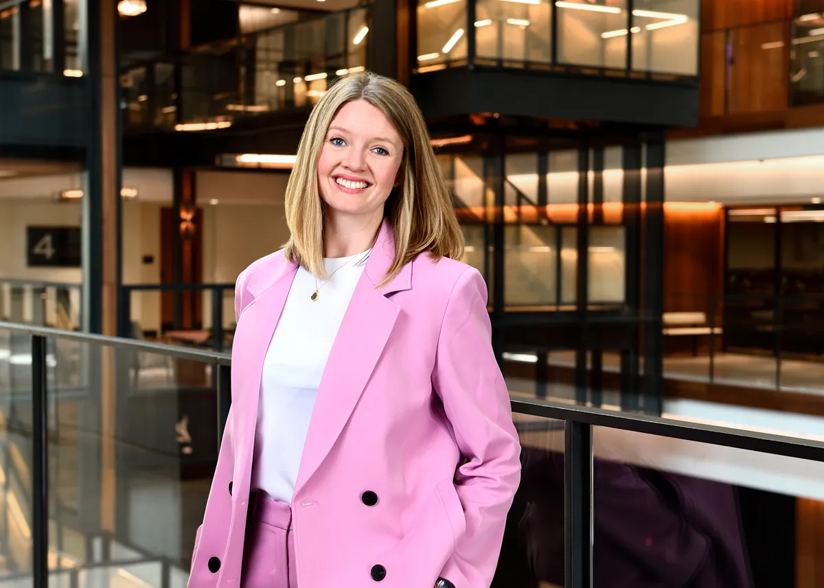 Portrait of Caitlin Berry, who wears a pink suit and stand in front of a balcony in a multi-level building.