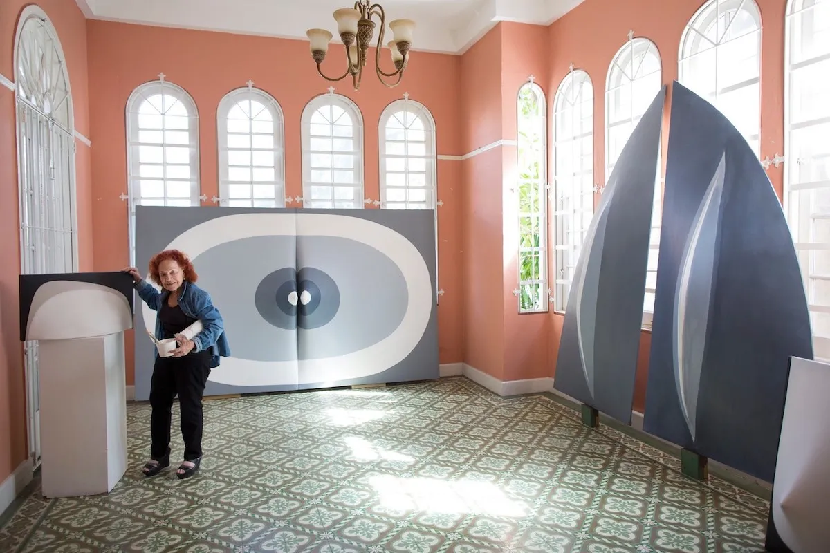 A woman with reddish hair standing amid giant abstract paintings in shades of grey. One leaned against a windowed wall features rings of white and grey that meet at a central cleavage.